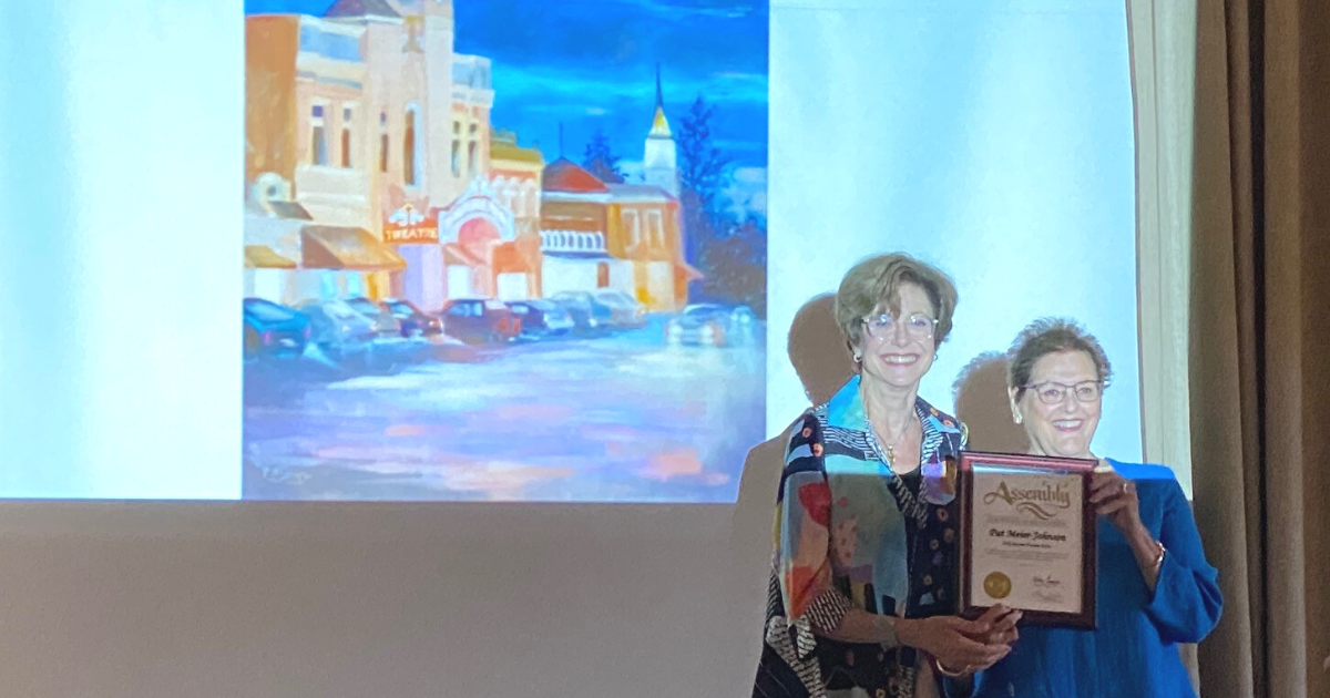 Two women holding a plaque and smiling in front of a projection of a painting of the Sebastiani Theatre in downtown Sonoma.