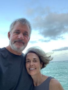 A man and woman standing close together smiling and looking at the camera with a body of water in the background.