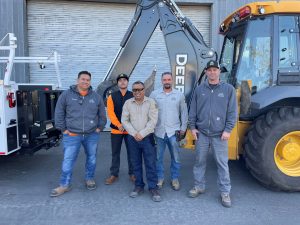 Five people standing in front of a backhoe and work truck.