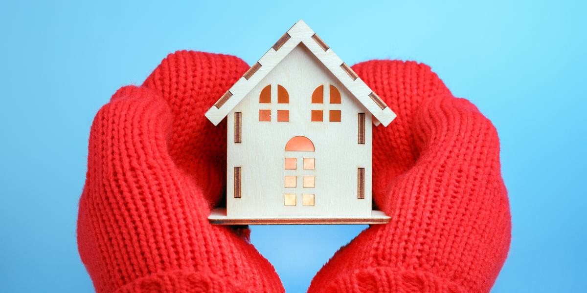 Photo of a two hands in bulky red mittens holding a miniature house made of cardboard.