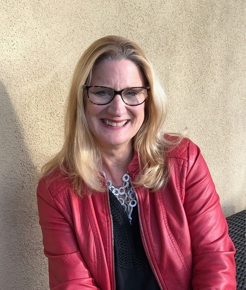 A woman sitting in sunshine with blond hair and glasses , wearing a red jacket and smiling at the camera.