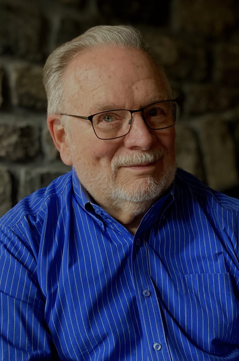 A man wearing glasses and wearing a blue striped button down shirt smiling at the camera with a stone wall in the background.