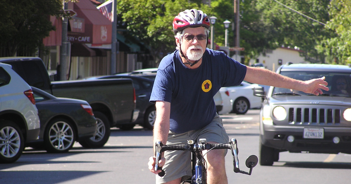 Man on a bike stopped at a stop sign with his hand outstreached to indicate that he is turning with a car in the background.