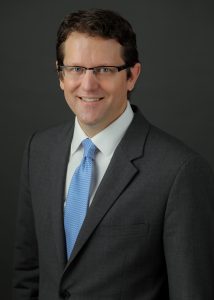 A man dressed in a suit and blue tie with glasses.