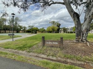 A park with grass and basketball court and play structure in the background with a sign that reads, Olsen Park.