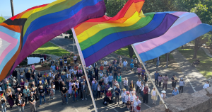 The progressive, tranditional and transgender pride flags flying in the wind on a balcolny with a crowd of people in the background.