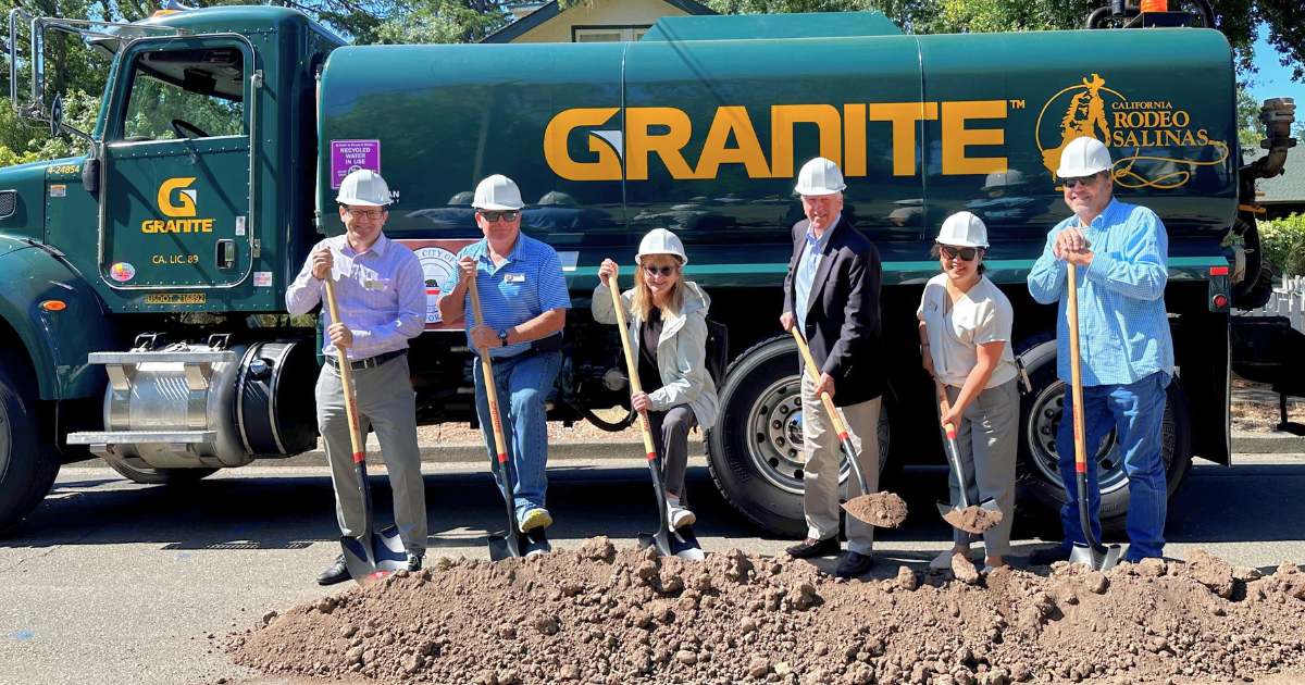 A photo of 6 people with shovels and hardhats in front of a large water truck shoveling dirt.