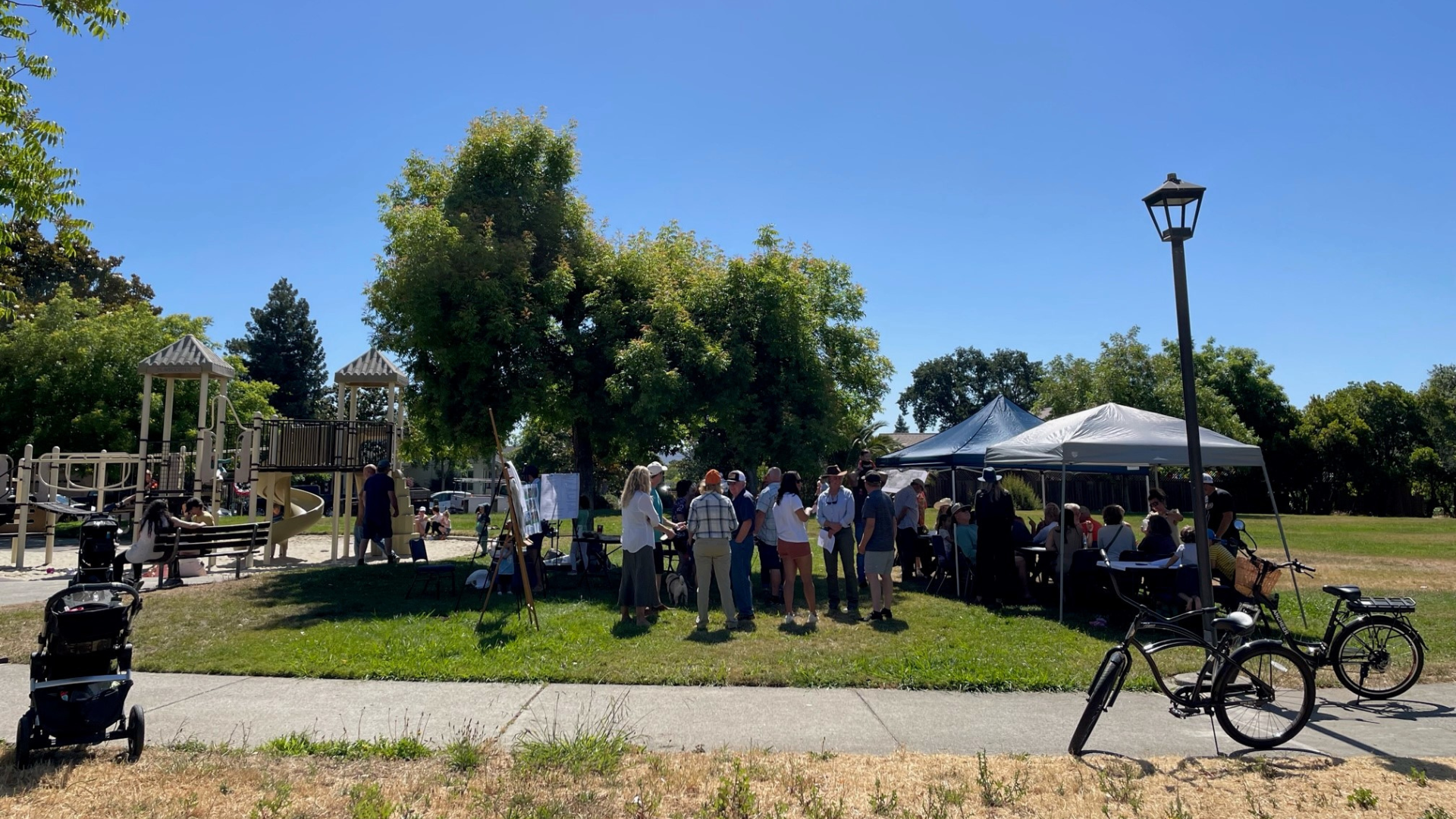 Photo of people gathered at a park with bikes and a stroller in the foreground.