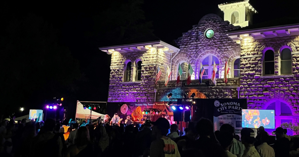 Sonoma City Hall at night with a stage and band playing and a crowd watching, the building is lit up in purple lights.