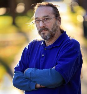 A photo of a man with glasses and wearing a blue button down shirt with his arms crossed in front of him and out of focus natural background.