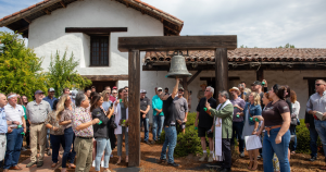 A crowd of people watching a man ring a large bell outside of the Sonoma Mission.