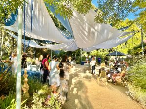 A lush garden with sun shades with a woman addressing a group of people.