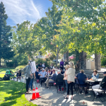 A group of people sitting at tables in a park looking at an easel.