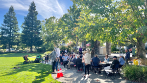 A group of people sitting at tables in a park looking at an easel.