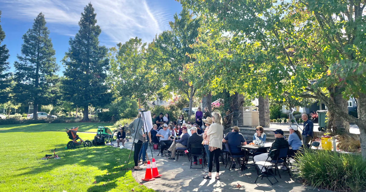 A group of people sitting at tables in a park looking at an easel.