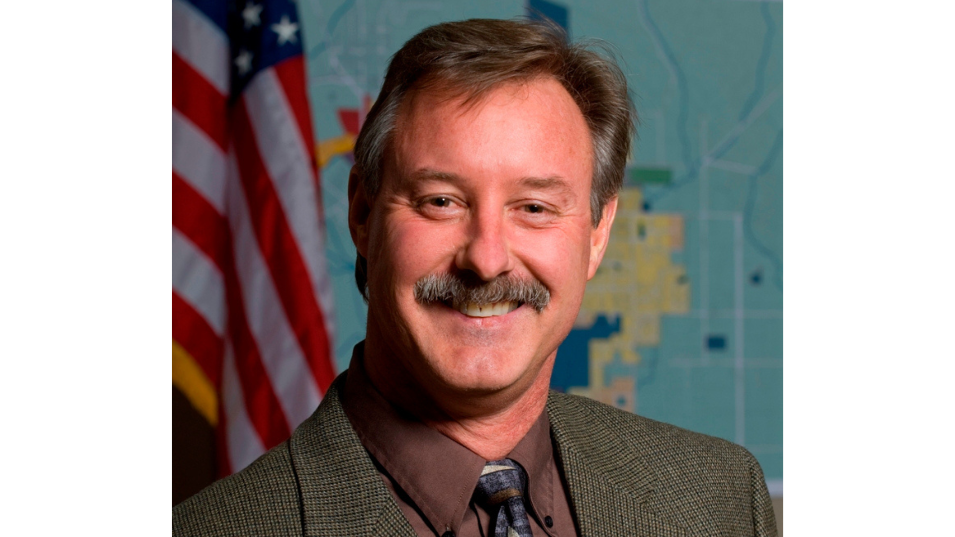 A photo of Wayne Wirick, who is smiling and positioned in front of an American Flag and a map of the City of Sonoma.