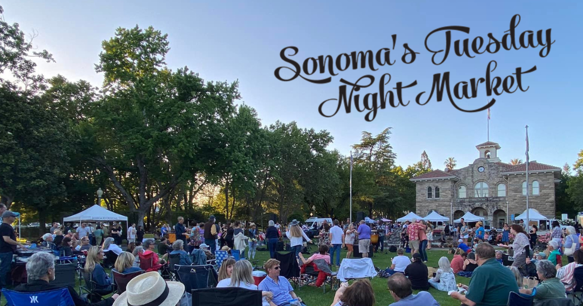 A photo of people picnicking and socializing on a large lawn with farmers market stalls and Sonoma City Hall in the background.