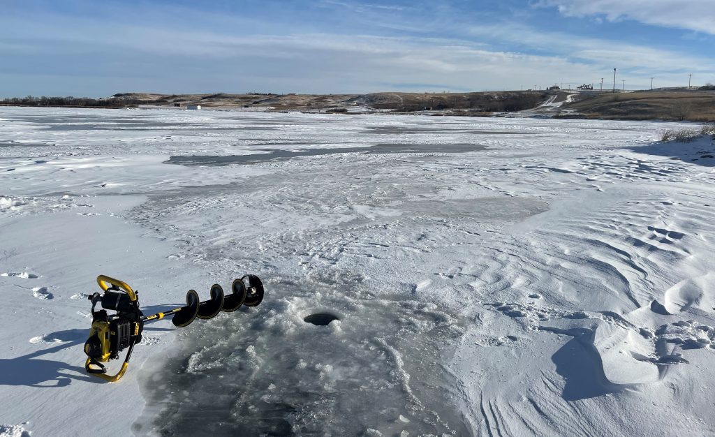 Ice auger laying next to a freshly drilled hole in an ice and snow covered lake.