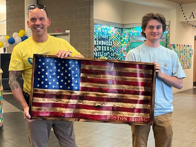 Der Ressourcenbeauftragte der Schule, Mike Chase, nimmt ein unerwartetes, aber passendes Geschenk der Kindness Week entgegen, eine ERSTAUNLICHE Boston Strong-Flagge, die geschnitzt wurde