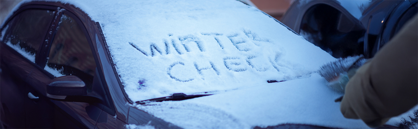 The front of a car covered in snow with the words 'winter check' written in the snow on the front window

