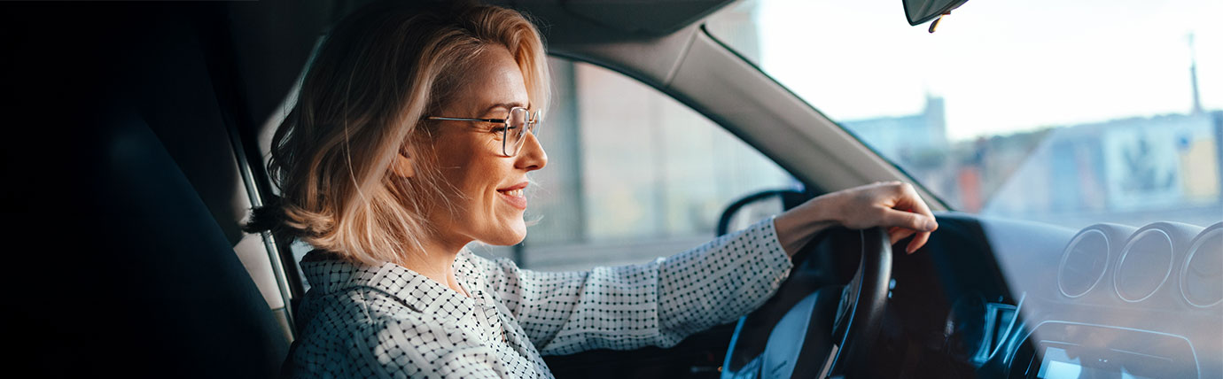 A blonde woman wearing glasses smiling while driving a car