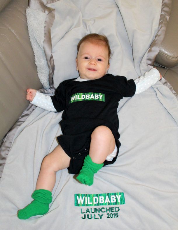 Photo of a baby in a onesie with "Wildbaby" across the front on a blanket with "Launched July 2015" embroidered on it.