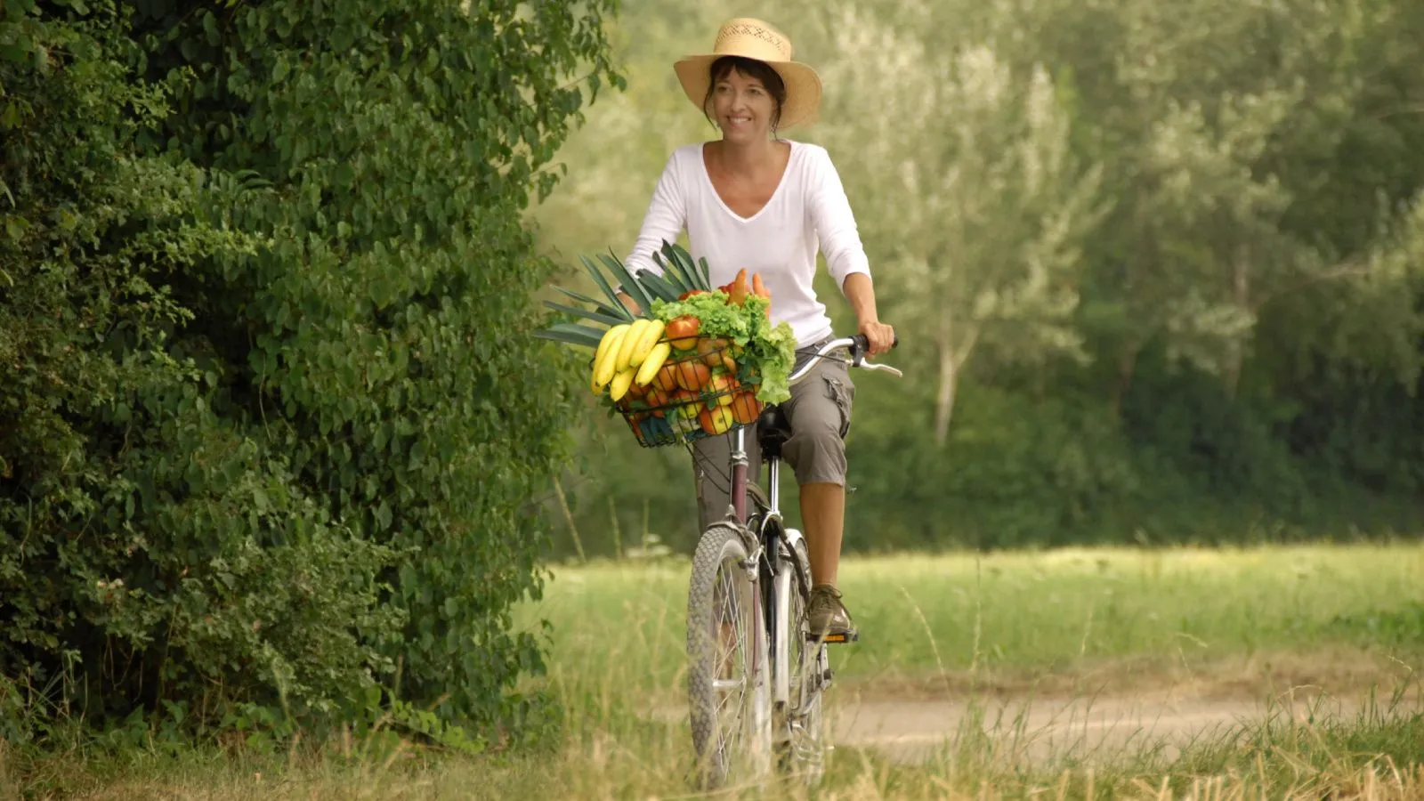 Frau fährt im Grünen auf dem Velo. im Korb hat sie Obst und Gemüse.