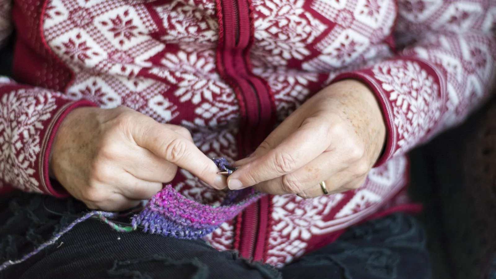 Hände und Arme einer alten Frau, sie trägt eine weiss-rot gemusterte Wolljacke und strickt