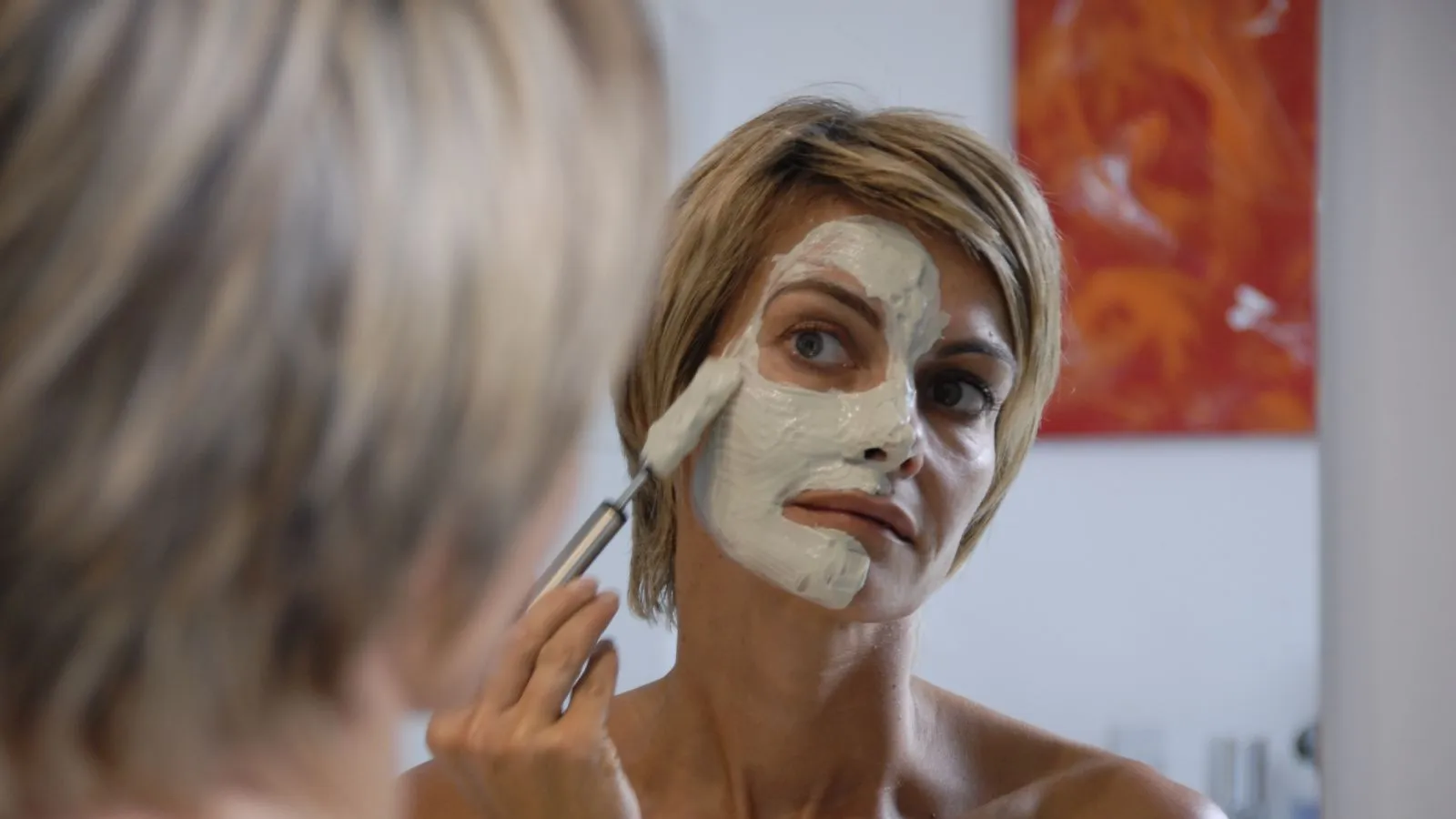 Frau mit blonden kurzen Haaren trägt mit einem Pinsel eine weisse Paste auf ihr Gesicht auf