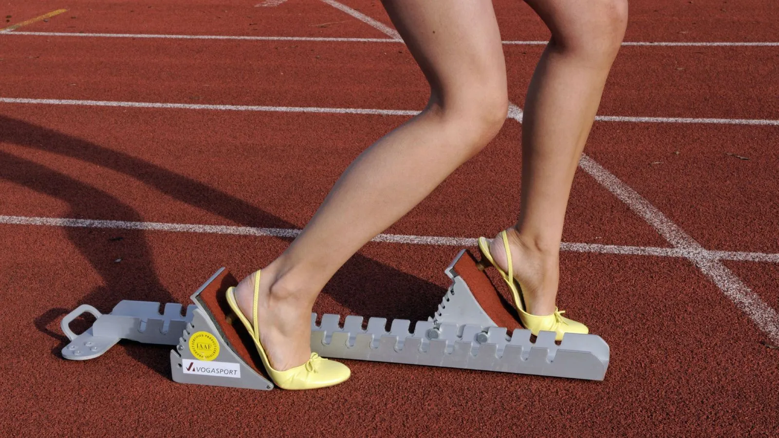 Frau mit nackten Beinen und gelben Stöckelschuhen auf einem Sportplatz. Nur die Beine sind zu sehen.