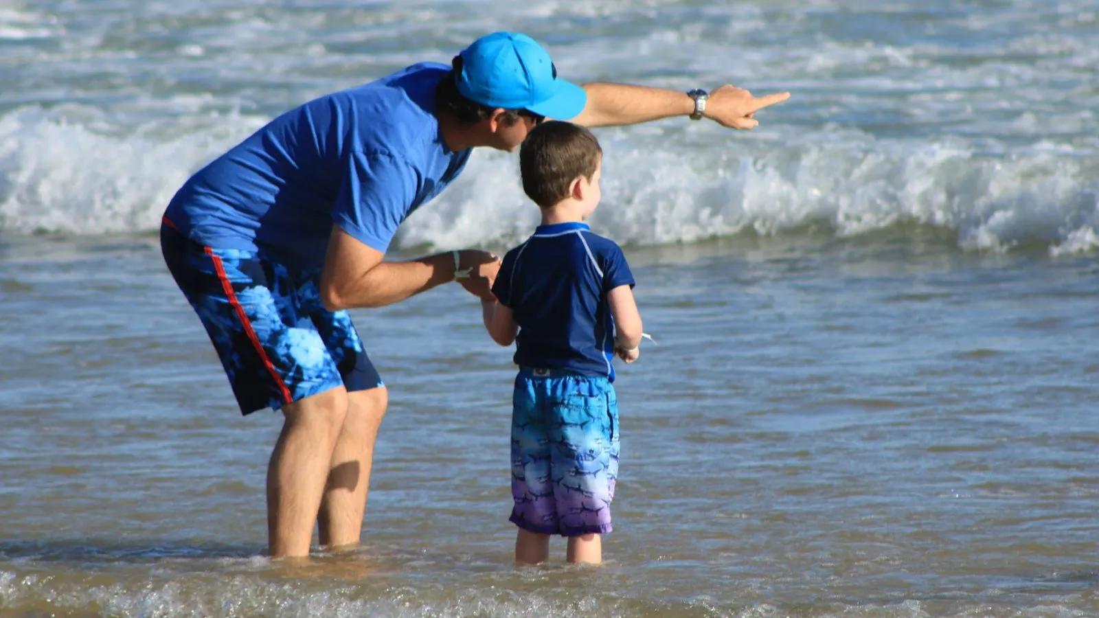 Ein Vater mit T-Shirt und Sonnenhut steht mit seinem Jungen im Wasser am Strand.