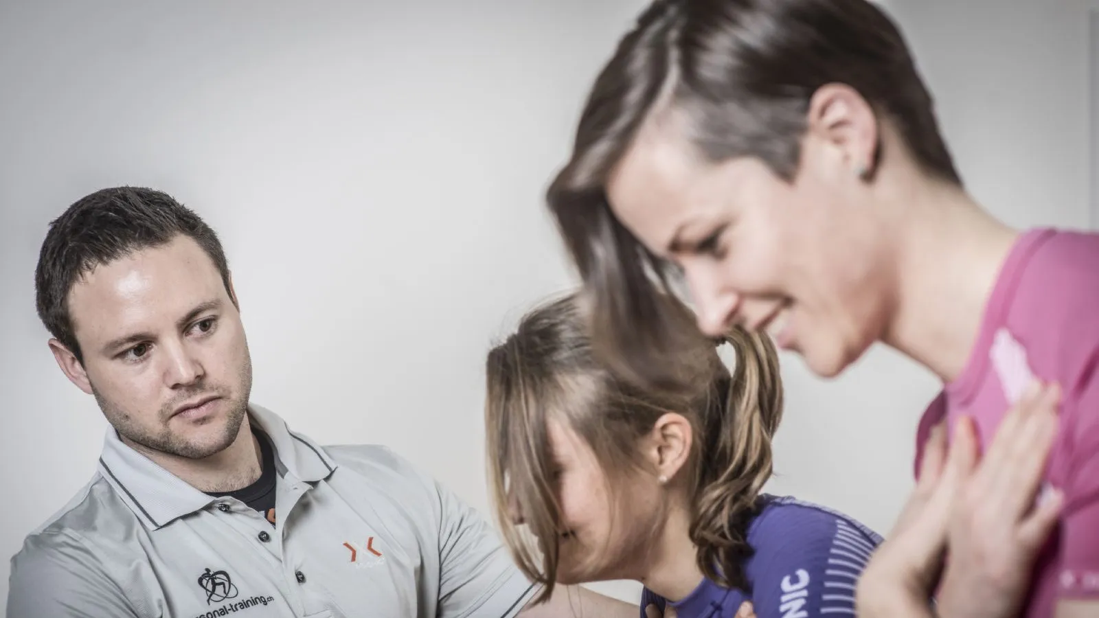 Zwei junge Sportlerinnen, sichtbar sind Kopf und Oberkörper üben unter der Aufsicht des Fitnesstrainers Kniebeugen.