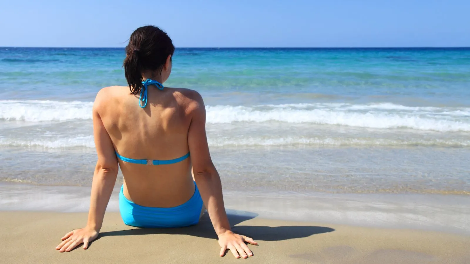 Sonnenschutz, Sonne, Meer, Strand: Junge Frau in blauem Bikini sitzt am Strand. Wir sehen sie von hinten.