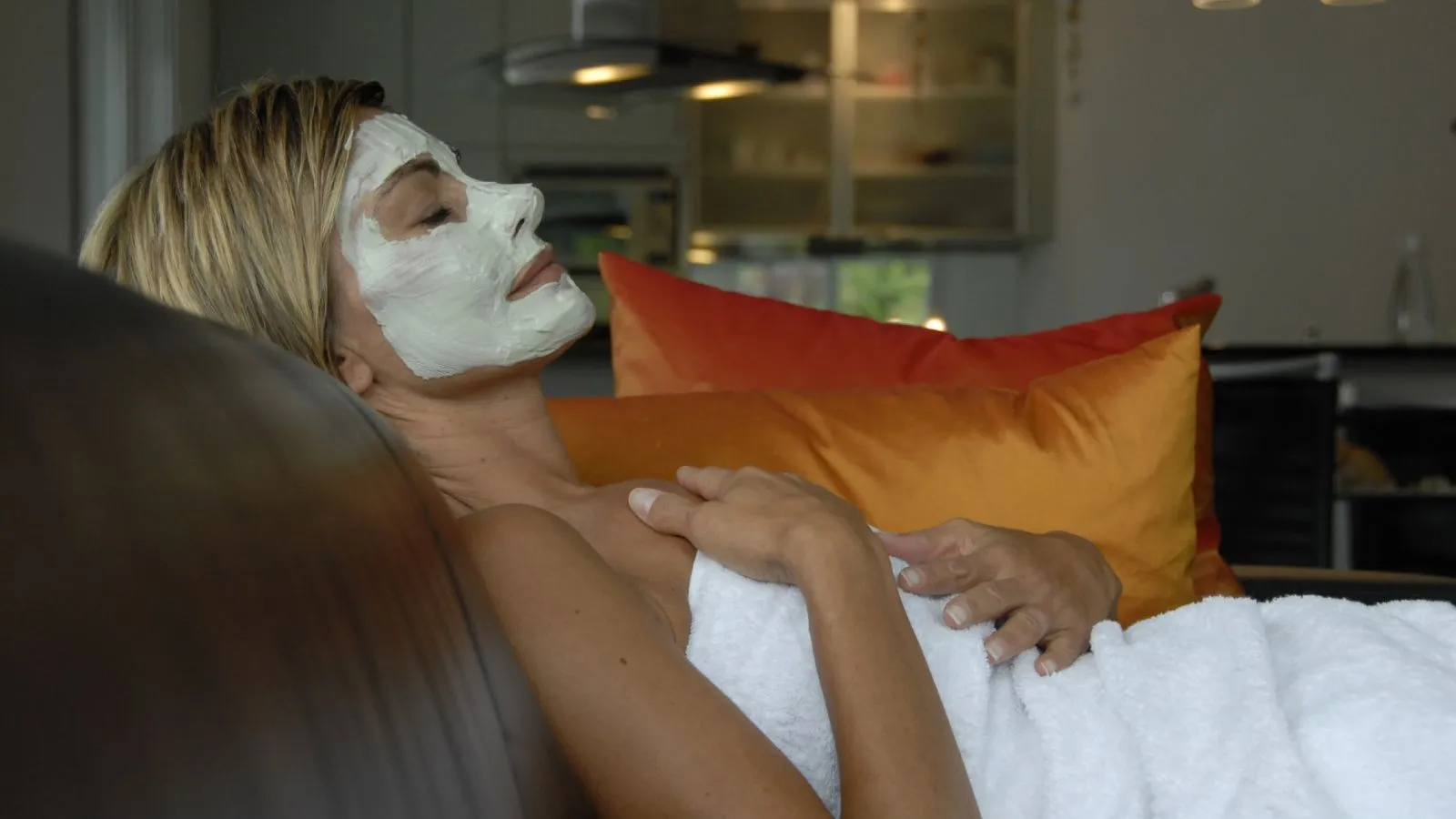 Frau mit kurzen blonden Haaren liegt, in ein weisses Frotteetuch gewickelt, auf dem Sofa, auf ihrem Gesicht eine weisse Gesichtsmaske