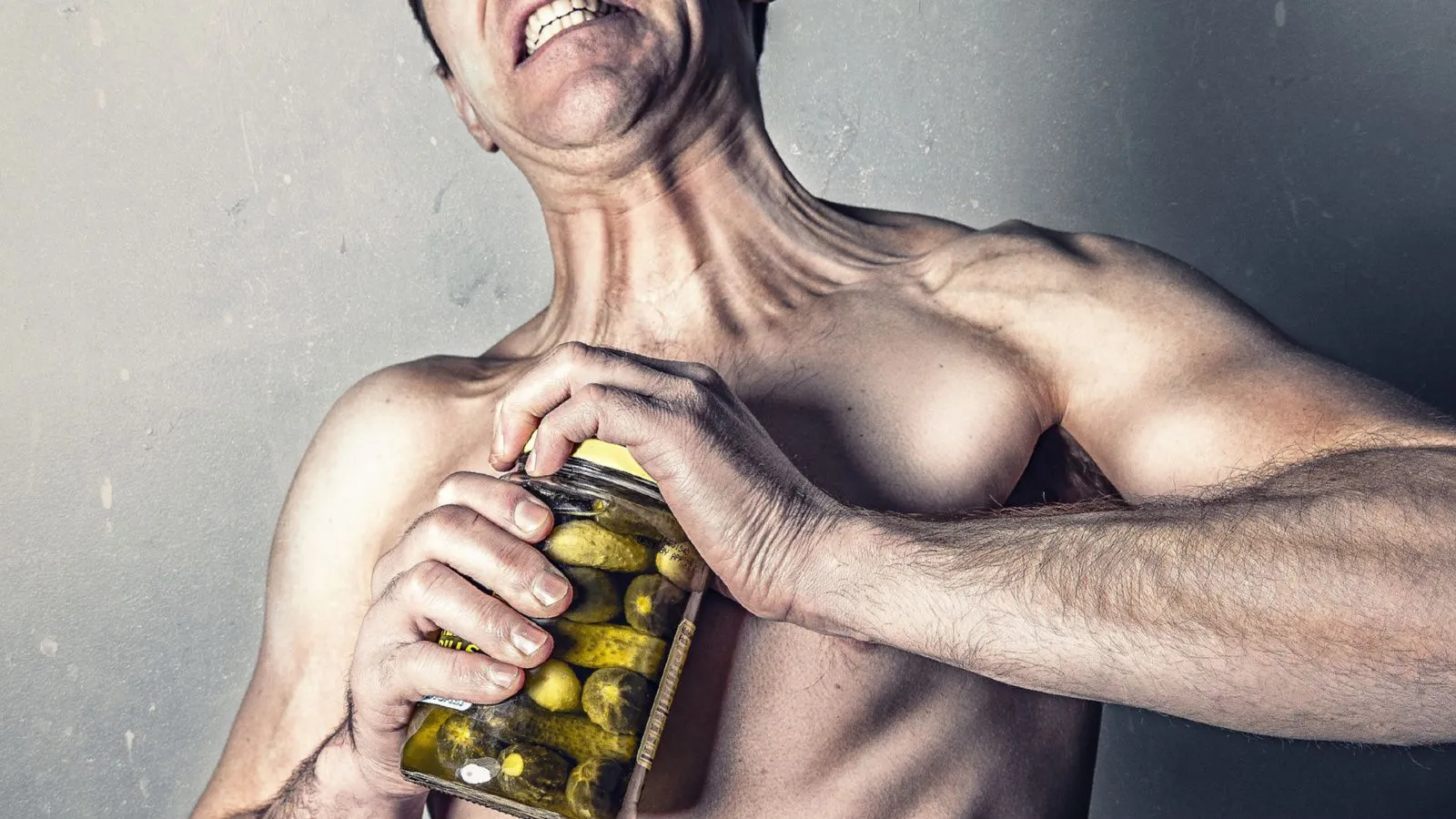 Mann mit nacktem Oberkörper versucht krampfhaft, ein Glas mit Essiggurken zu öffnen. Seine Muskeln zeichen sich ab