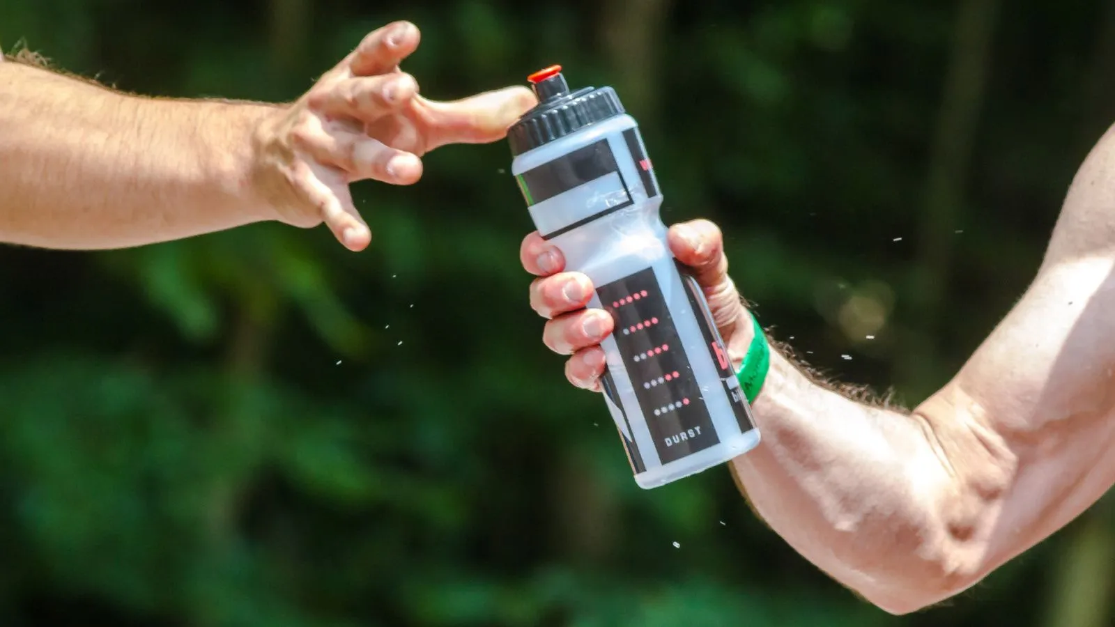 Zwei Männerarme, einer hält in der Hand eine Trinkflasche aus Plastik, die der andere sich gerade greifen möchte