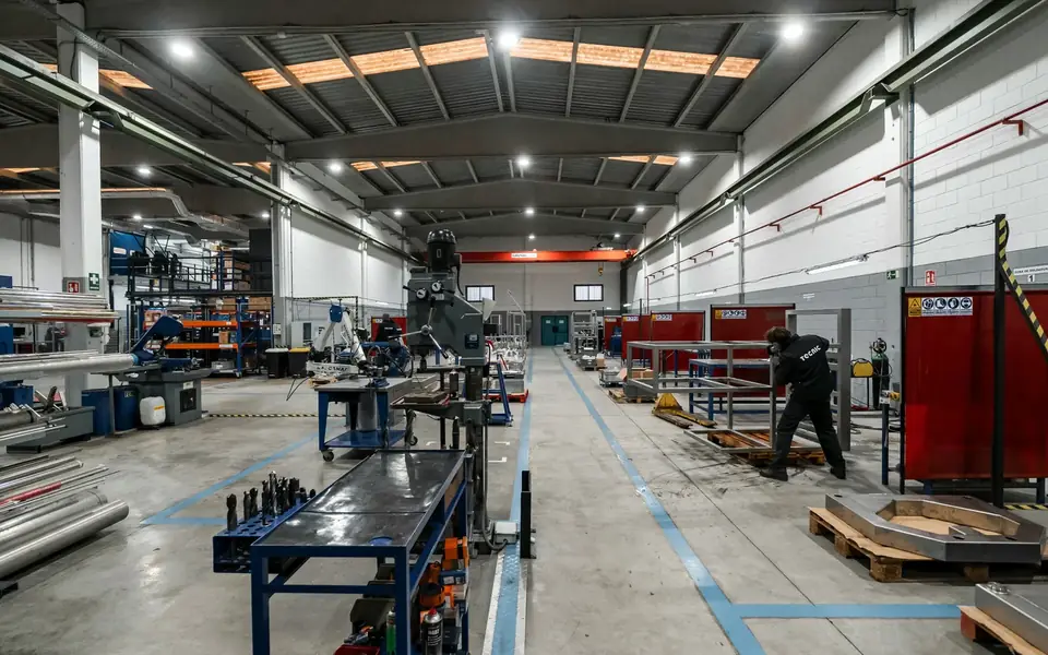 Atelier d'une PME industrielle : rayonnages de profilés métalliques, établis et un opérateur au travail.