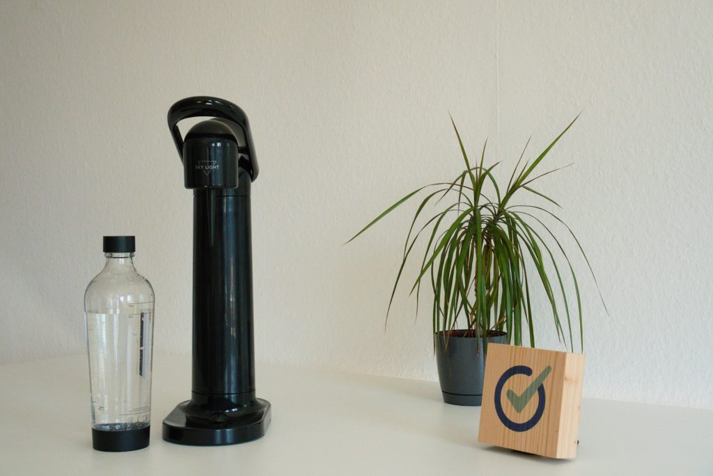 Sky Light Wassersprudler neben PET-Flasche neben Pflanze und Holzblock mit testit-Logo