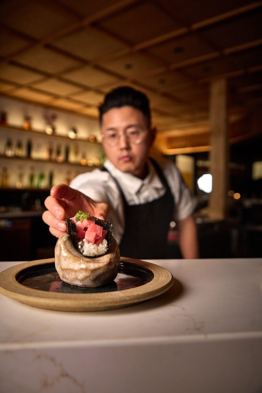 A chef in a white shirt and blue apron places a handroll onto a stone shaped plate.