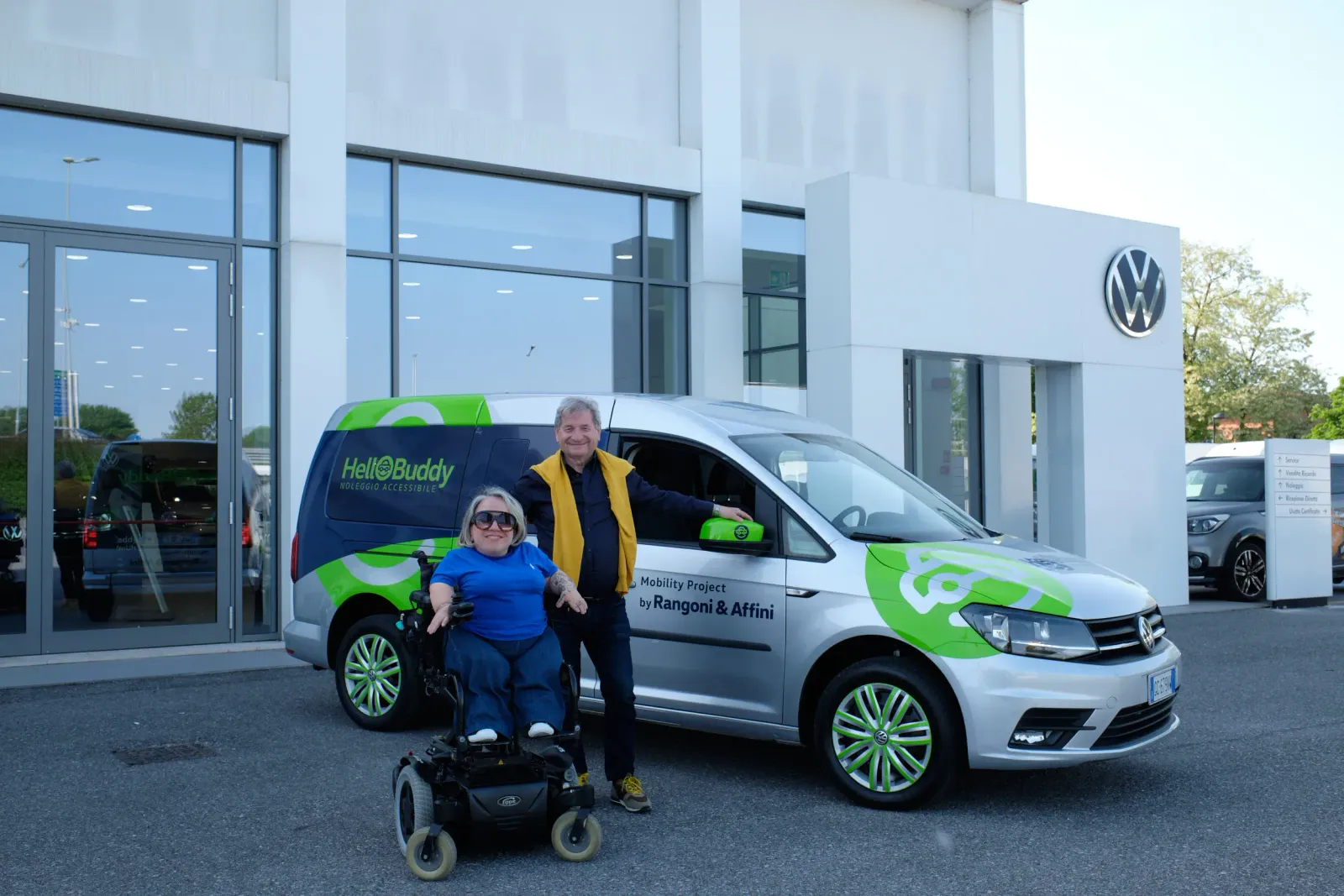 Una donna in sedia a rotelle e un uomo posano accanto a un furgone "HelloBuddy Noleggio Accessibile" di fronte a una concessionaria Volkswagen.