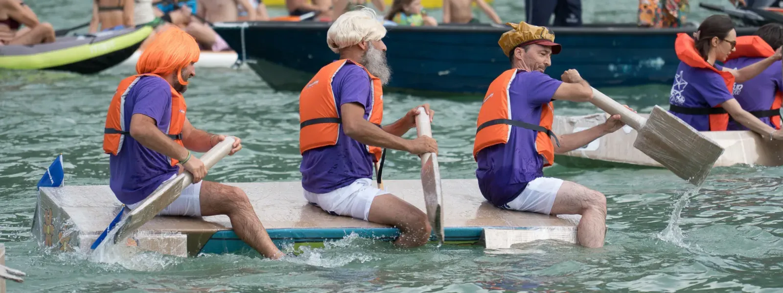 Una gara di barche di cartone con partecipanti vestiti in modo eccentrico, che remano vigorosamente in acqua.