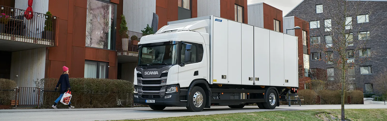 Un camion Scania è parcheggiato sul lato della strada mentre una donna cammina lungo il marciapiede.