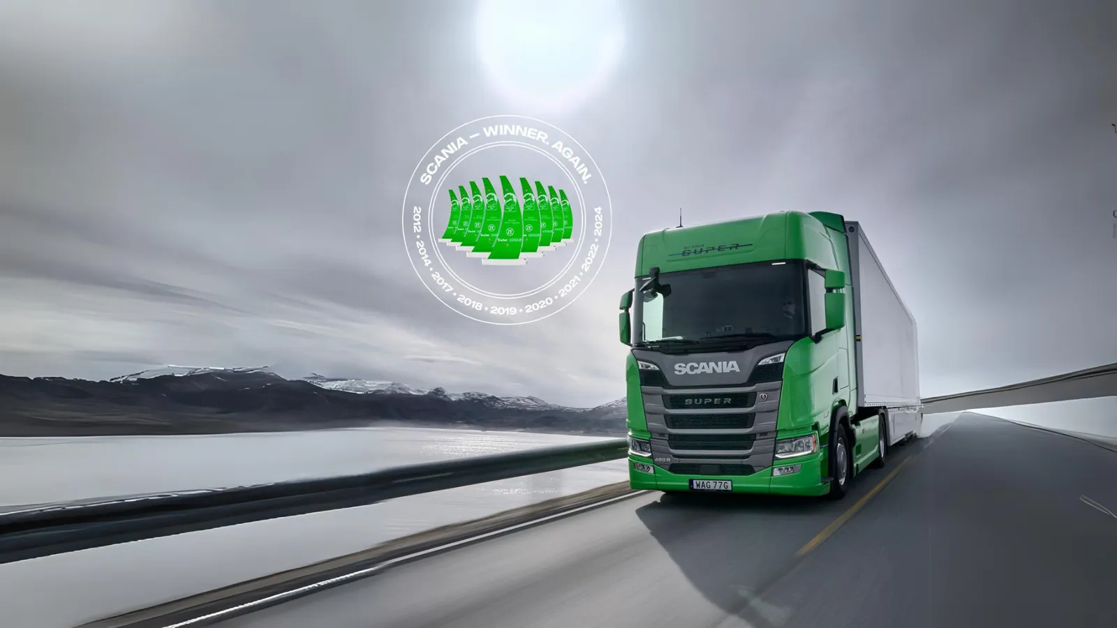 Un camion Scania Super verde guida su una strada con un lago e delle montagne innevate sullo sfondo.