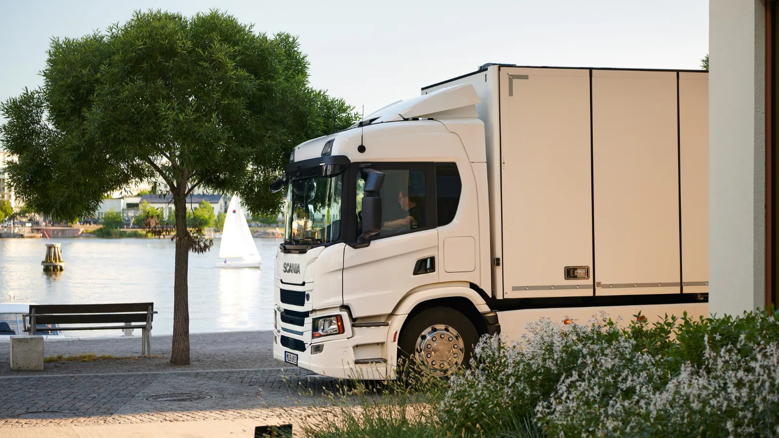 Un camion Scania bianco parcheggiato vicino a un lago con una barca a vela in lontananza.