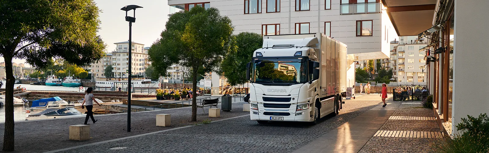 Una persona attraversa una strada acciottolata mentre un camion bianco passa accanto a ristoranti e bar affollati vicino al lungomare.