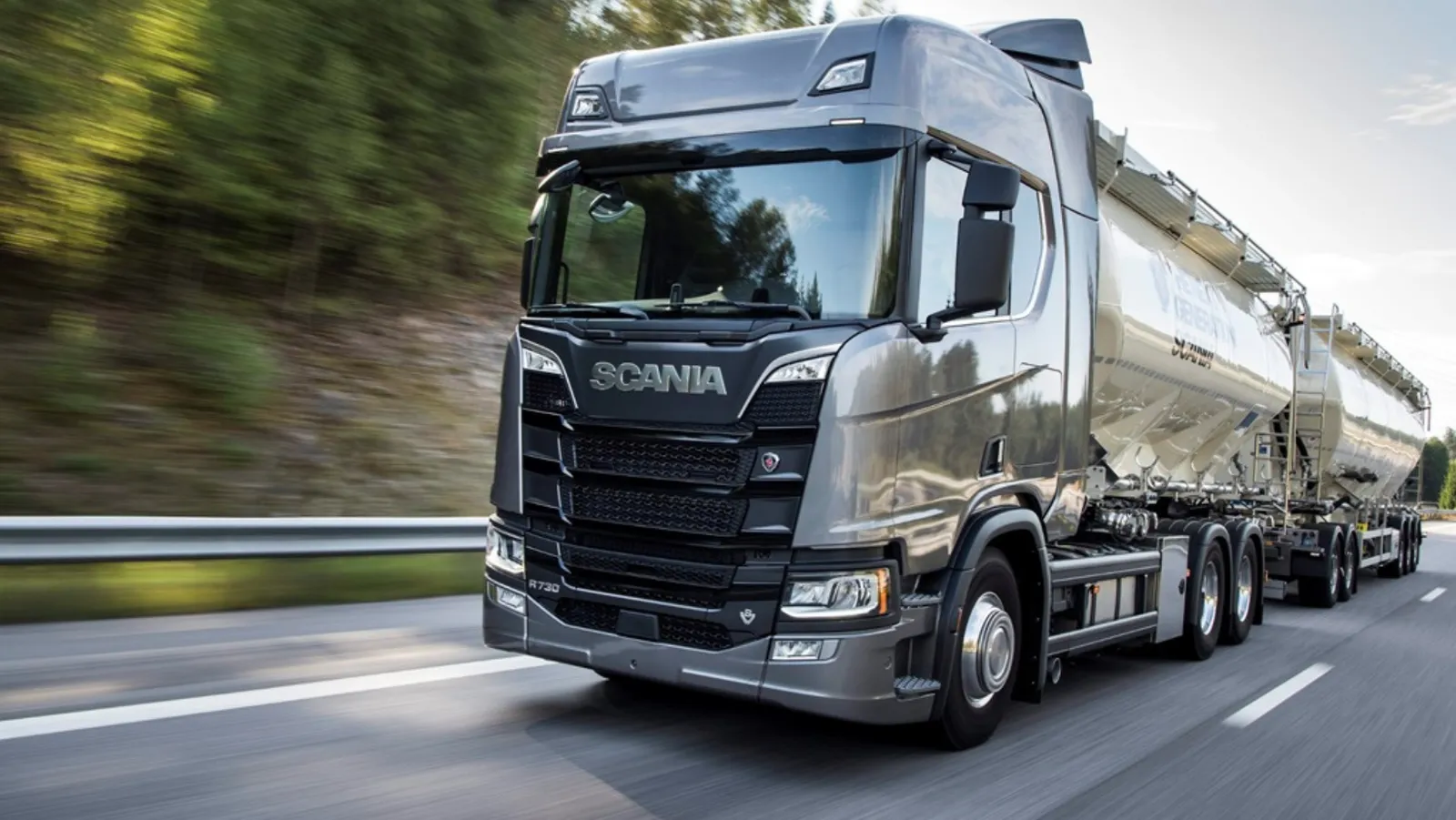 Un autocarro Scania grigio con rimorchio a silos in movimento su una strada.