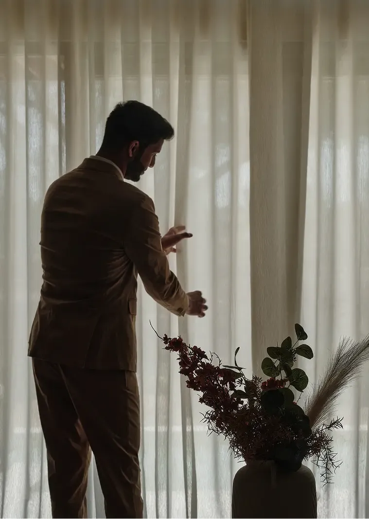 Man in a light brown suit adjusts sheer white curtains, silhouetted against the light.