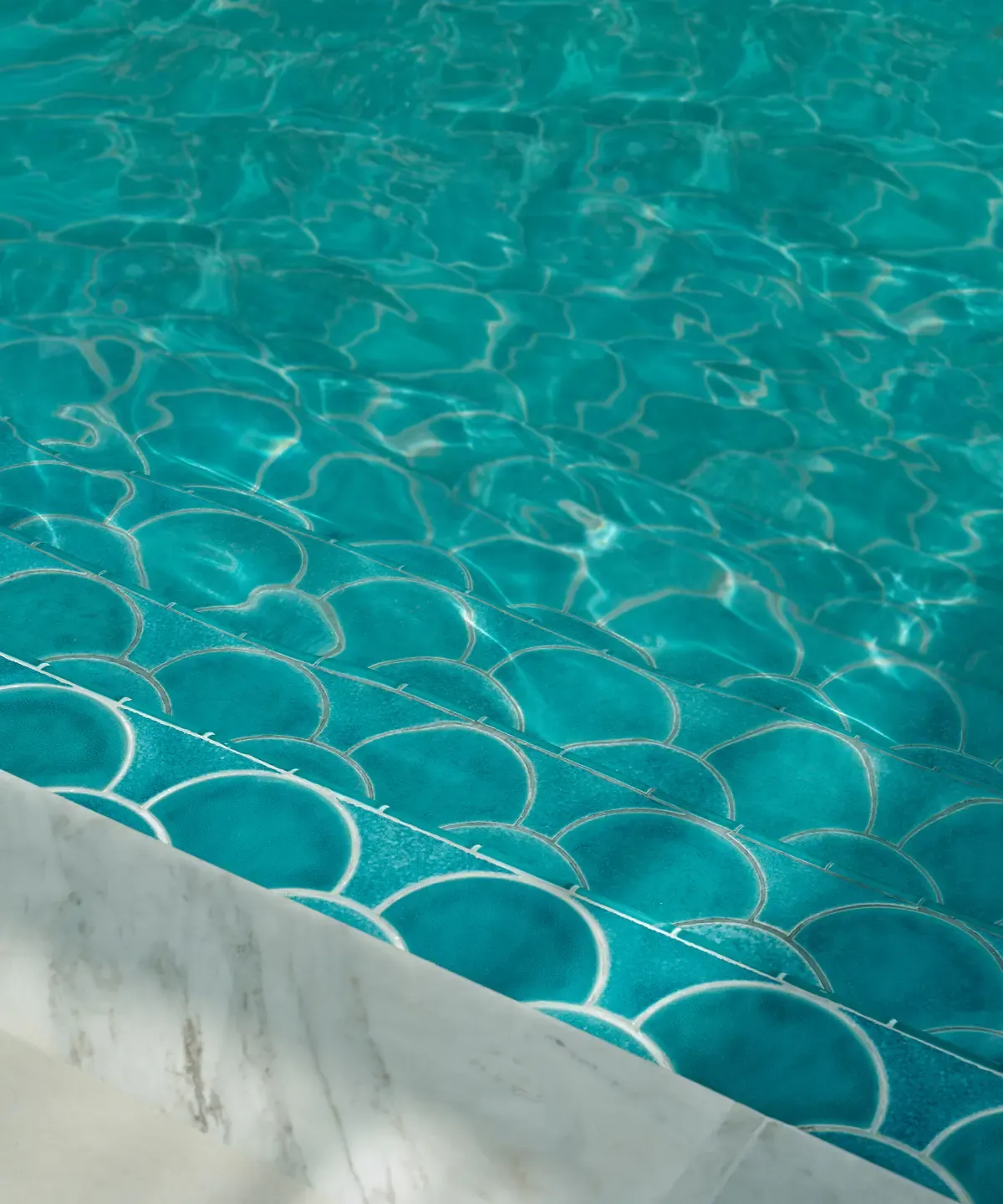 Clear turquoise pool water with sunlit ripples over light blue scale-patterned mosaic tiles.