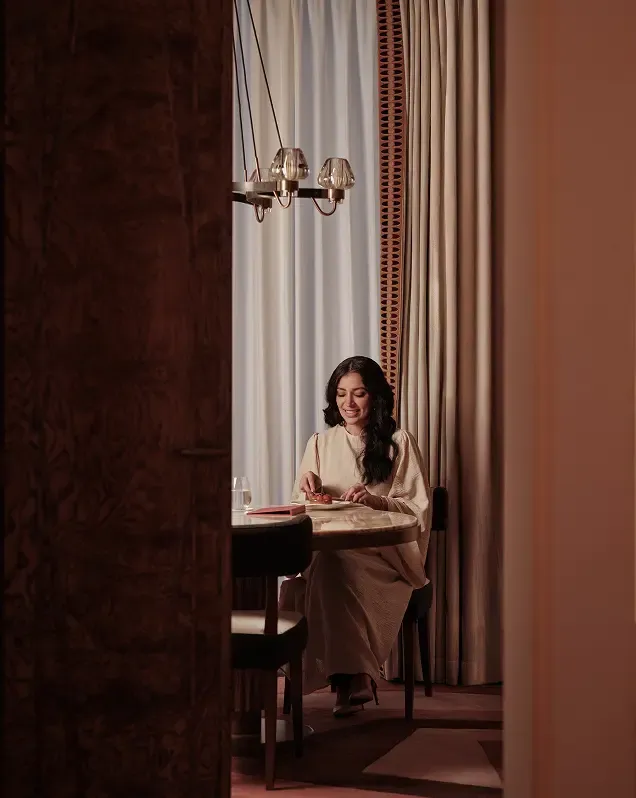 Smiling woman in a cream-colored dress sits at a wooden table, enjoying a meal indoors.