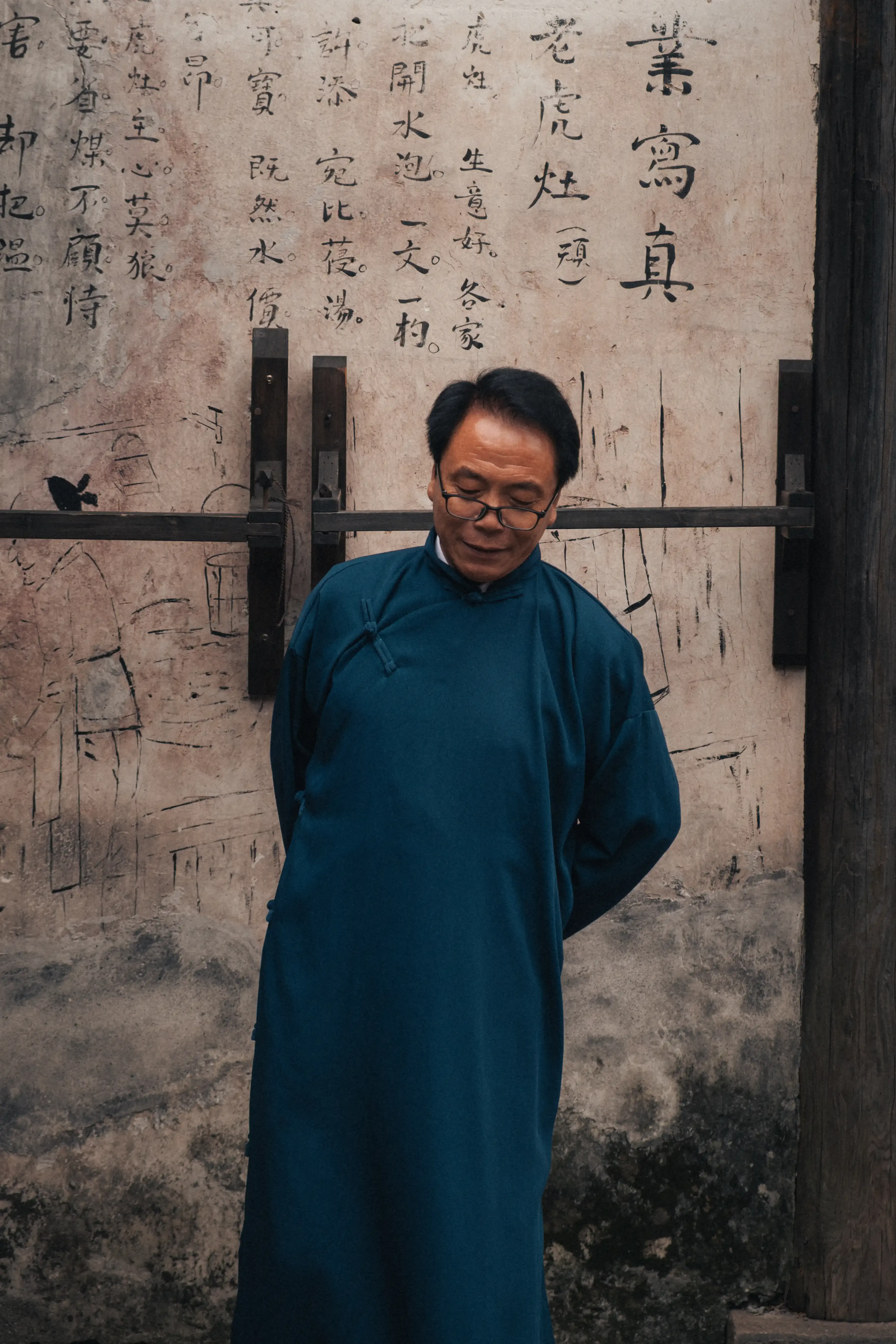 Portrait d'un homme âgé en longue tunique bleue traditionnelle debout devant un mur couvert de calligraphies chinoises anciennes
